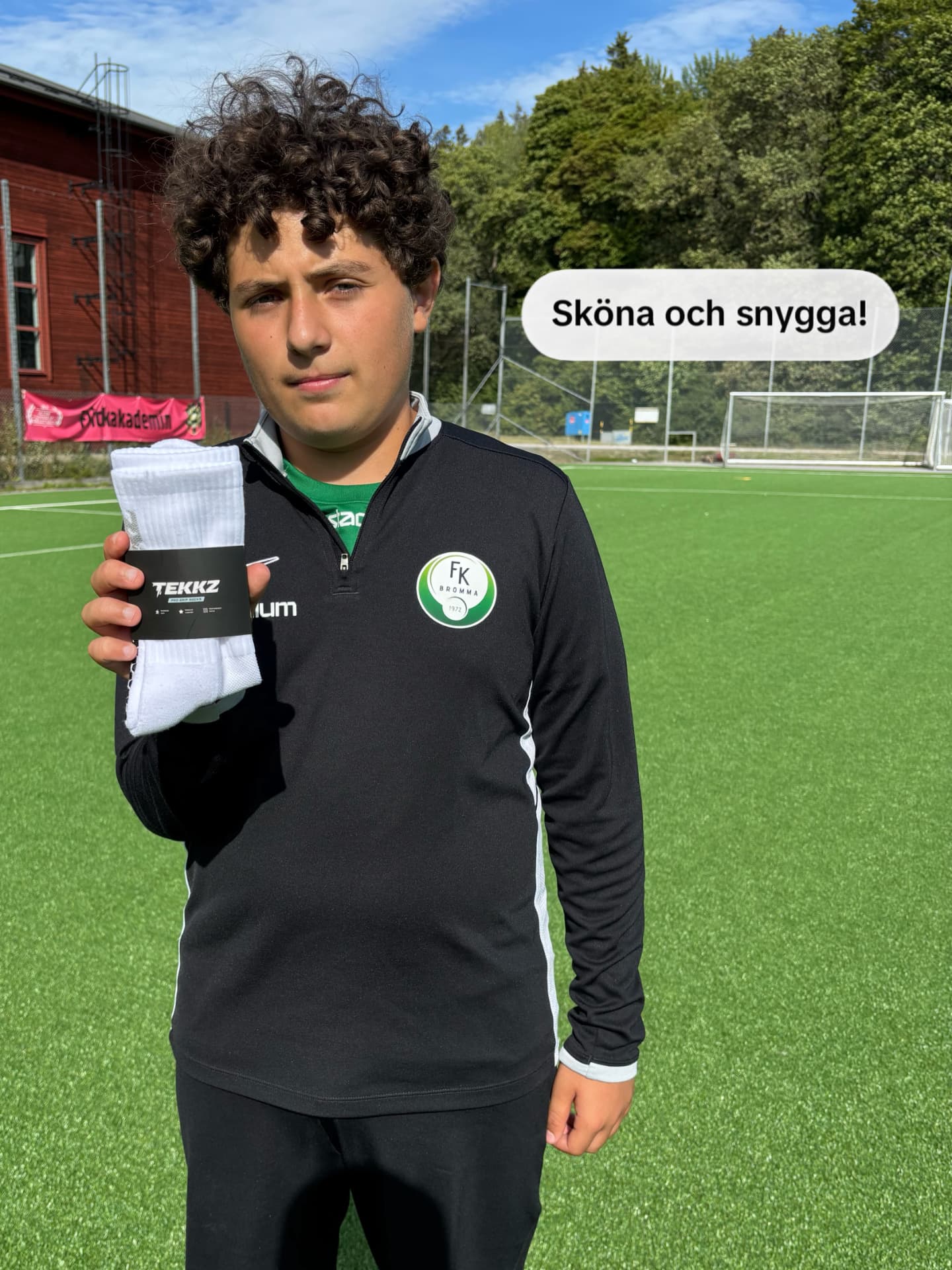 Ung spelare i svart TEKKZ-tröja håller upp ett par vita TEKKZ greppstrumpor på fotbollsplanen – "Sköna och snygga!"